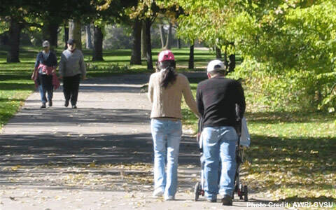 Two pairs of pedestrians walk on a long sidewalk through a treed park.